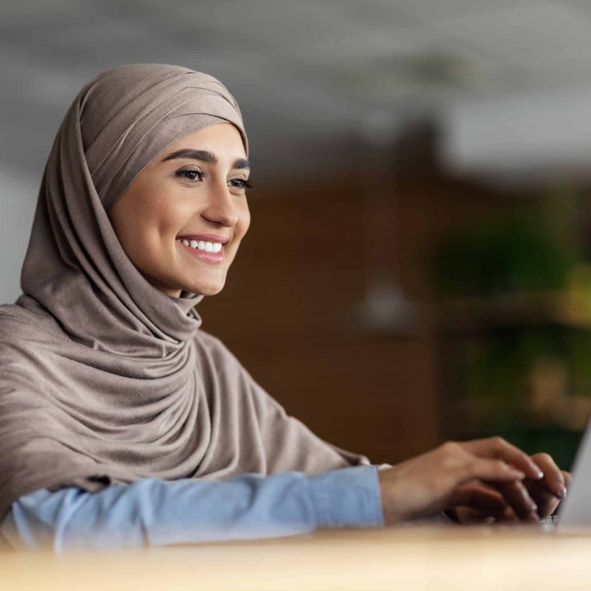 Cheerful arab lady in hijab working online at cafe, using laptop, copy space. Side view of smiling muslim woman freelancer typing on laptop, enjoying her new distant job, chatting with clients