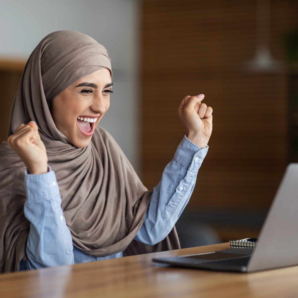 Emotional muslim woman in headscarf looking at laptop screen Emotional muslim woman in headscarf looking at laptop screen and raising hands up. Happy arab lady using laptop at cafe got good news, found nice job , empty space, career and job seeking concept
