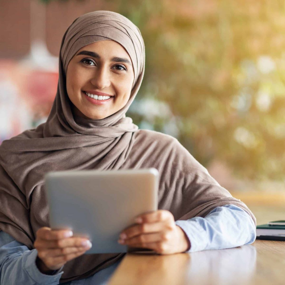 Happy girl in hijab with digital tablet smiling at camera, sitting at cafe alone, copy space