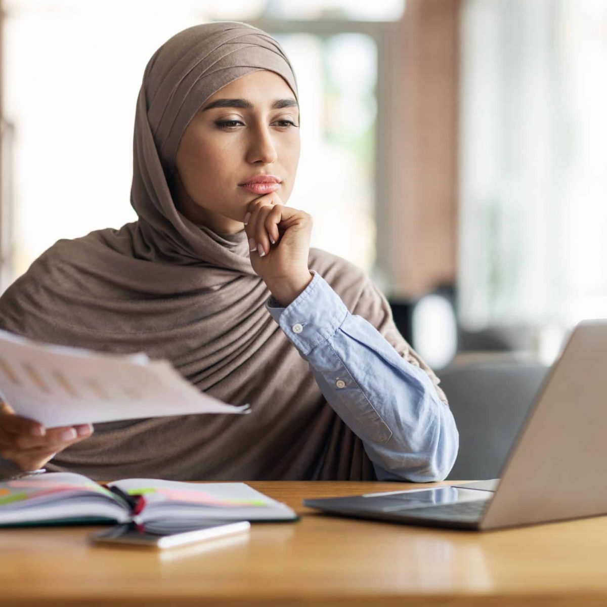 Pensive muslim woman manager working while sitting at cafe Pensive muslim woman manager working while sitting at cafe, using laptop and checking documents, copy space