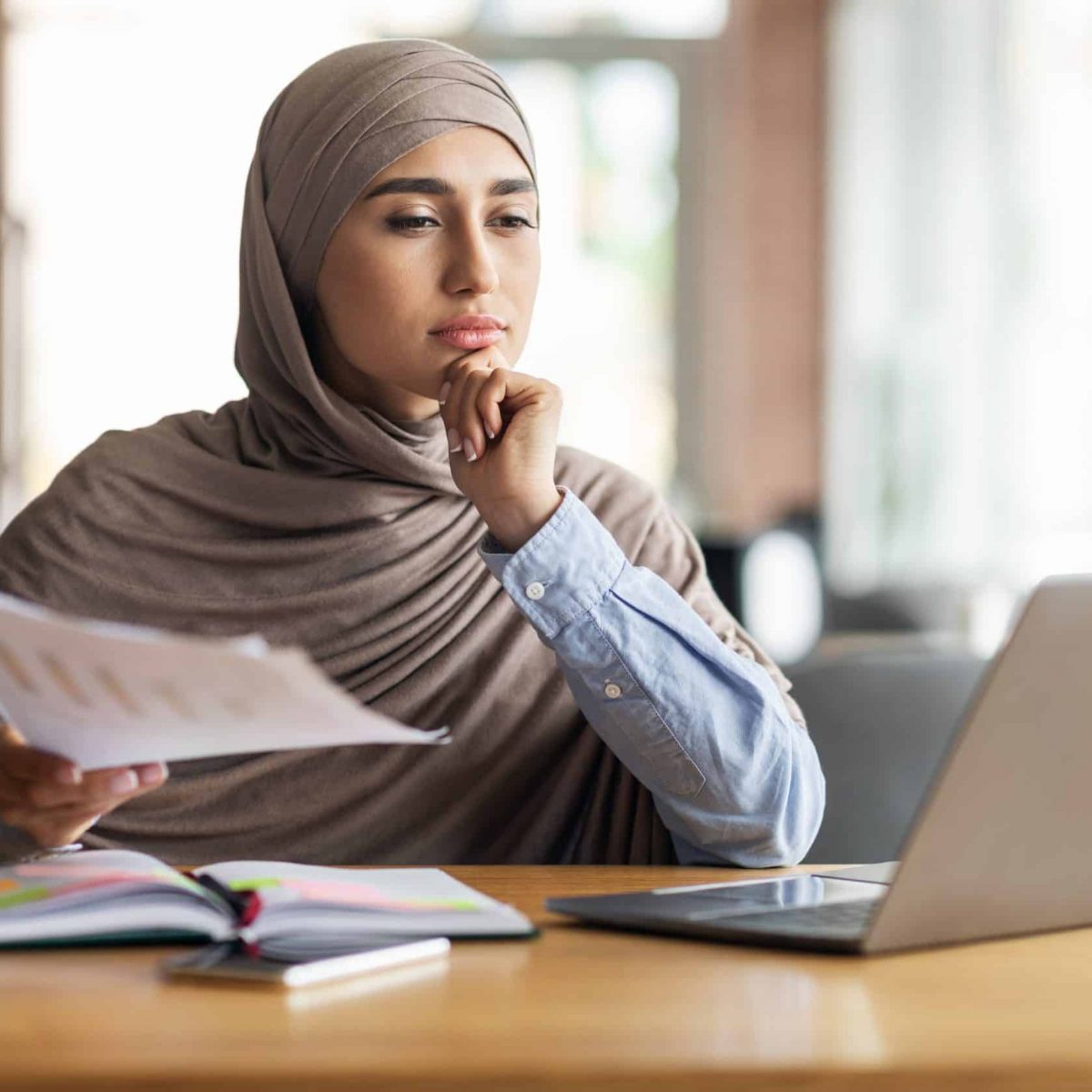 Pensive muslim woman manager working while sitting at cafe, using laptop and checking documents, copy space